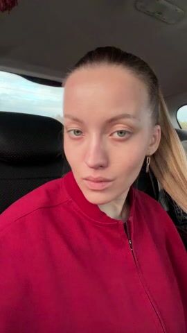 A light-skinned young woman with blonde hair in a ponytail sits in the backseat of a car. She is wearing a red bomber jacket and gold earrings. She looks at the camera and smiles slightly, and then closes her eyes, as if dozing off, before looking at the camera again. A brown tassel hangs from the car's ceiling.