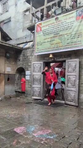 A woman wearing a red headscarf and a red and black salwar kameez walks barefoot across a wet, stone courtyard. She carries a blue plastic bag in her right hand. The courtyard is decorated with colorful chalk markings. To her left is a stone structure covered with a green and yellow cloth, possibly an idol or monument. In front of her, an open doorway leads into a building, revealing glimpses of people and religious iconography inside. A bell hangs above the doorway. A banner with text in a non-English script is displayed above the entrance. The architecture appears to be of Indian origin. The overall scene suggests a visit to a temple or religious site.