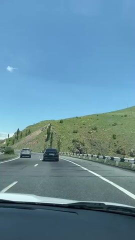 This is a first-person perspective video taken from inside a car driving on a highway. The car is in the right lane. In front of the car are two other vehicles, a black SUV and a white car. The road is a multi-lane highway with a clear blue sky and some white clouds overhead. To the right of the highway is a green, rocky hillside with some scattered trees. To the left of the highway, the landscape appears flatter and more developed with more cars visible in the distance. The video is stable and appears to be shot on a sunny day.