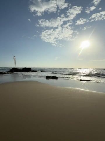 The scene shows a beach on a sunny day. The tide is coming in on the sandy shore. The sun is high in the sky with some scattered clouds. Footprints mark the wet sand. In the distance, there are some rocks and a flag.