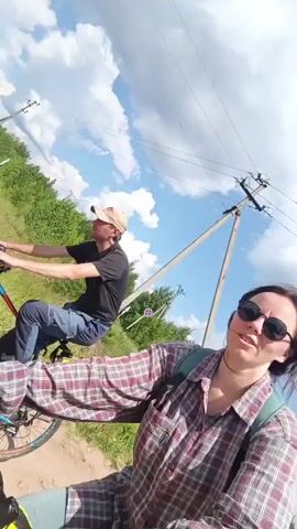 Two people are riding bicycles on a dirt road with grassy and wooded areas on the sides. A power line runs parallel to the road. The sky is blue with white clouds. The person in the foreground is a woman wearing a plaid shirt, sunglasses, and a backpack. The person behind her is a man wearing a black t-shirt and a baseball cap. Both are looking forward and appear to be cycling at a moderate pace. The video is filmed from the perspective of the woman in the foreground.
