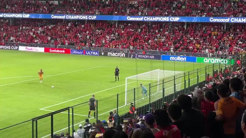 The video shows a soccer player in an orange jersey taking a penalty kick at a crowded stadium. The crowd is primarily dressed in red, and a referee stands nearby. After the player kicks the ball, the camera shakes as the crowd erupts in cheers, presumably because the player scored. The backdrop shows advertisements for various sponsors like Scotiabank, Michelob Ultra, and Riyadh Air. The event is identified as the CONCACAF Champions Cup.