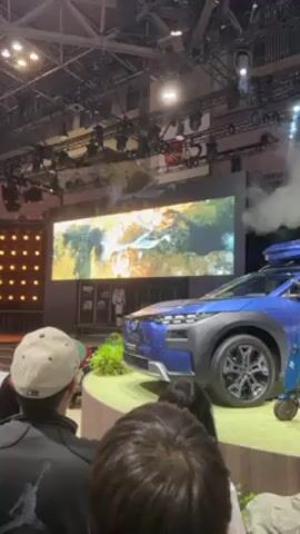 The video shows a car presentation on a rotating stage. The car is blue and sits on a grassy platform surrounded by onlookers. There is a large screen behind the car, displaying nature scenes. Vapor effects are present, contributing to a dramatic reveal. A matching scooter is positioned next to the car. The environment appears to be a large event hall, likely a car show, as indicated by the lighting, stage setup, and audience. A person in the front is wearing a grey hat with a logo on it.