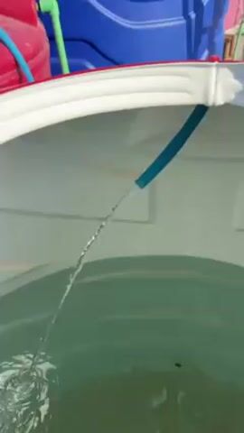 A blue hose pours water into a large, white, circular container that already contains water. The containers surrounding the white container include a red one and a blue one, and there is a green hose connected to the red container.