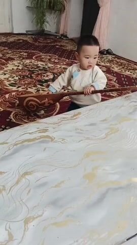 A young child with dark hair is seen on a patterned rug inside a room. The child, dressed in a cream-colored shirt, is crouched behind a large piece of fabric that is white with gold swirls. The child is holding a stringed instrument that is resting partially on the rug and partially on the fabric. The instrument appears to be made of wood. A large potted plant is visible in the background on the left. In the background, a figure is partially visible near a doorway with light pink curtains. The child is looking down at the fabric and touching it with their fingers.