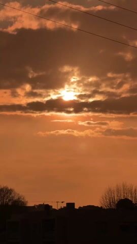 The video shows a sky with an orange hue. The sun is visible through clouds, creating a dramatic effect. Power lines cut across the top of the frame. Silhouetted buildings and trees are visible at the bottom of the frame.