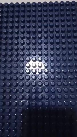 A blue baseplate with raised circular studs is shown from a top-down perspective. The lighting creates highlights and shadows on the surface, suggesting it is made of plastic and may be used for building blocks.