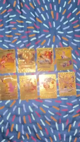 A medium shot shows eight gold-colored, foil Pokemon cards on a blanket. The blanket is blue with a confetti-like pattern made of dashes of orange, pink, and white. Each card has an image of a different Pokemon on it. The camera zooms in to show the details of the cards.
