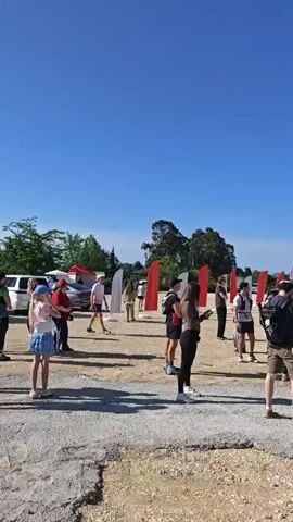 A daytime, low-angle, medium shot shows people standing around a sandy, gravelly area under a clear, bright blue sky. A red tent is on the right, and a blue and white inflatable archway is on the left. Several people are standing around chatting and looking at things. Most of the people are in casual clothing, with some wearing athletic wear. A small dog with a leash is among the people. There is a sign on the red tent and some flags next to the archway. In the background are trees and other greenery.