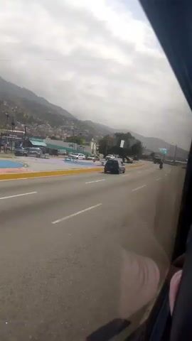 The video depicts a view from inside a vehicle moving along a multi-lane highway. On the left side of the road, there's a colorful plaza with benches and several parked vehicles. Mountains are visible in the background under a cloudy sky. Cars and a motorcycle are driving along the highway.