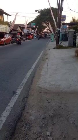 A street scene with several motorcycles, cars, and buildings. Flags are hanging over the street. The weather appears to be clear and sunny.
