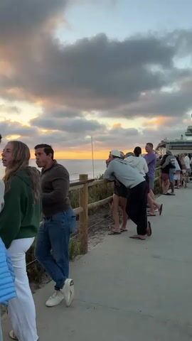 A group of people are standing on a paved walkway next to a wooden railing. They are looking out at the ocean and the sky, which is filled with dramatic clouds lit by a setting sun. The foreground features a man in a brown shirt and blue jeans, leaning against the railing. Behind him, several other individuals are gathered, some appearing to converse. The environment is coastal, with vegetation visible along the edge of the walkway. The overall atmosphere suggests a shared appreciation of the sunset.