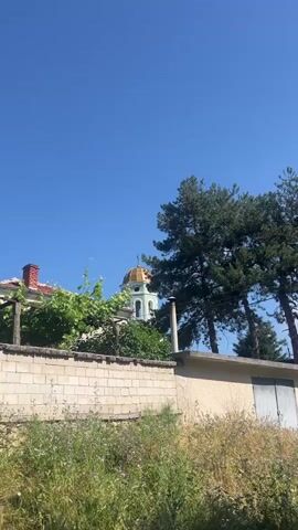 The video shows a close-up shot of a church steeple with a golden dome against a clear blue sky. Lush green trees frame the steeple on the right side of the image, and leafy vines grow on the left. In the foreground, a low brick wall separates the viewer from the church. The wall is partially covered with tall, dry grass and wildflowers. To the right of the church, a portion of a building with a beige wall and a closed garage door is visible. A white pipe extends vertically from the beige wall, possibly a chimney or vent. The sun is shining, casting a bright light on the scene, suggesting it is daytime.