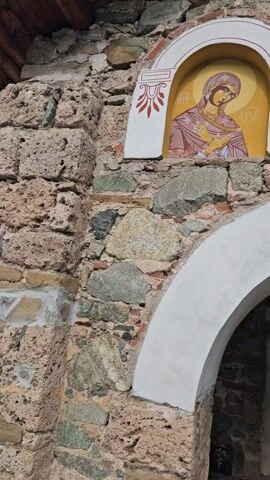 The video shows a close-up, upward-tilting shot of the exterior of a stone building. Above a white arched entryway are three painted icons. The leftmost icon depicts the Virgin Mary, the central one shows Jesus Christ holding a book, and the rightmost icon presents a bearded male saint. Underneath the arch of the entryway, a small, framed portrait of a bearded man hangs above a black door. The building’s facade is constructed from a mix of stones and brick. The icons are set within white frames with decorative red and white elements. The style of the paintings suggests a Byzantine or Orthodox Christian theme. The camera slowly moves up to reveal the icons.