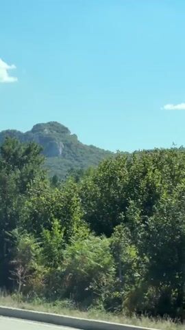 A video shot from a moving vehicle captures a picturesque view of a large, rounded mountain covered in greenery, under a clear blue sky with a few white clouds. The foreground is dominated by lush trees and vegetation, with a road divider visible in the lower portion of the frame. The camera appears to be in motion, suggesting a scenic drive.