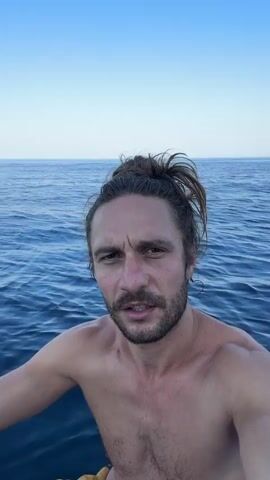 A man with his hair tied up and a mustache is on a body of water. He is shirtless and looking at the camera. The water is blue and calm. In the background, there are hills and trees. The sky is clear and blue.