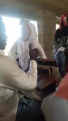Here is a description of the video. 

The video shows a person holding a baby indoors. The baby is wearing a white outfit and a white head covering. The person holding the baby is wearing a white shirt and dark pants. There are other people in the room, including one wearing a black and white striped shirt and jeans, and one wearing a headscarf. The room appears to be a classroom or meeting room, with wooden desks and benches. There is a window in the background providing light. The camera angle is slightly low, looking up at the person holding the baby. The focus is on the baby and the person holding them. The audio captures conversation.