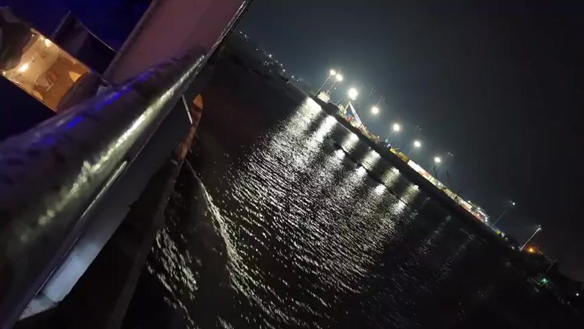 This video captures a nighttime scene from a bridge overlooking a body of water. The water's surface is rippled, reflecting the lights from streetlamps lining the bridge or a distant shoreline. In the foreground, a metal railing with a blue light strip runs diagonally across the frame, obscuring part of the view. The overall atmosphere is dark and serene, with the illuminated water creating a sense of movement and depth. No people or specific objects other than the railing and lights are clearly discernible.