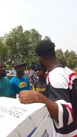 Here is a concise description of the video:

The video shows a group of people standing outdoors, possibly discussing something related to the sign they are standing next to. The sign is for "CJAY AUTOSWHEEL ENTERPRISES" and lists "OUR SERVICES."  The man closest to the camera is wearing a black and white shirt with red accents and is speaking, gesturing with his hands. The others are listening to him, their expressions vary from neutral to somewhat concerned. One of the men is wearing a "Glovo" shirt. The setting appears to be a sunny day on a street or parking lot with trees in the background. In the background, there is a parked car, people, and motorcycles can be seen.