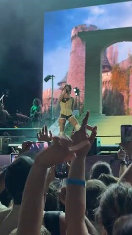 The video shows a live music performance in Segrate, Italy. The central subject is a female singer on stage, wearing a corset-like top and shorts, performing in front of a large screen displaying a castle backdrop. She is holding a microphone and appears to be interacting with the audience. To her left, two other individuals are visible on stage, one at a keyboard and another with a guitar.

In the foreground, a crowd of people are enthusiastically watching the performance. Many are holding up their mobile phones to record the event, with their hands raised in the air. The lighting appears to be stage lighting, with green and white spotlights illuminating the stage. The overall atmosphere suggests a lively concert or festival. The time of day is likely evening or night, as the stage lighting is prominent and the background is dark. The emotion conveyed by the crowd is one of excitement and engagement with the performance. There is no visible text in the media file.