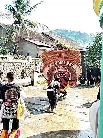 The video shows a traditional Indonesian performance in a rural setting. Dancers in traditional attire are moving in a circle, carrying props including a large, arched structure with the words "ANOM JOYO TALUN" written on it. Other participants carry shields and wear animal-like masks. The performance takes place on a wet concrete surface in what appears to be a village street. Houses with tiled roofs and lush greenery are visible in the background, suggesting a tropical or subtropical environment. There is a crowd of onlookers watching the performance, and the video is shot from within the crowd, partially obstructed by a green pole and an overhanging concrete structure.