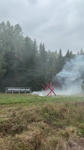 A reenactment of a historical battle is taking place in a grassy field with trees in the background. Smoke is billowing around a red anti-tank obstacle. Several signs with Russian text are visible in the background. People dressed in historical military uniforms are engaged in combat, with some holding rifles. There are also onlookers present, some of whom are filming the event. The sky is overcast.
