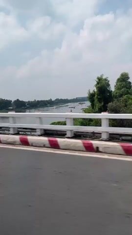 The video shows a view from a vehicle traveling across a bridge. The camera is positioned low, focusing on the side barrier of the bridge, which is painted white with red and white striped accents along the base. Beyond the barrier, a wide river is visible, with green vegetation lining its banks. A building is distantly visible on the horizon. The sky is partly cloudy. The road surface is dark gray asphalt. The vehicle moves forward.