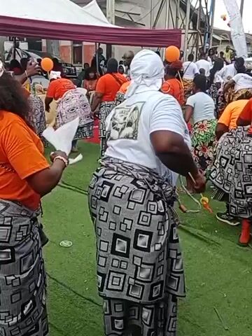 The video shows a group of people, predominantly women, gathered outdoors. Many of the women are wearing orange tops and traditional patterned wraps around their waists. Some are also wearing headscarves. The ground appears to be covered in green artificial turf. In the background, there are tents and buildings, suggesting an event or celebration is taking place. Some of the individuals are holding orange balloons and appear to be dancing or participating in an activity. The overall atmosphere seems festive.