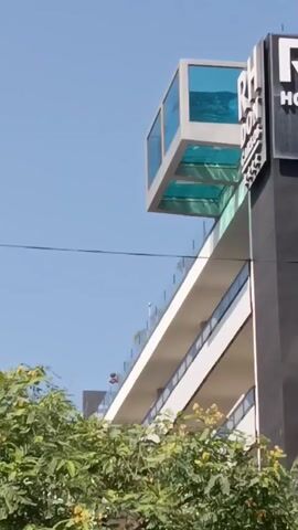 The video shows a building with a glass-enclosed swimming pool extending from the top. The pool is filled with blue water and appears to be suspended over the edge of the building. Below the pool, there are multiple levels of balconies with glass railings. In the distance, on one of the lower balconies, a person can be seen standing. The building has a black facade with a white sign that reads "RH HOTEL". Green foliage is visible in the foreground, obscuring the lower part of the building. The sky is clear and blue.