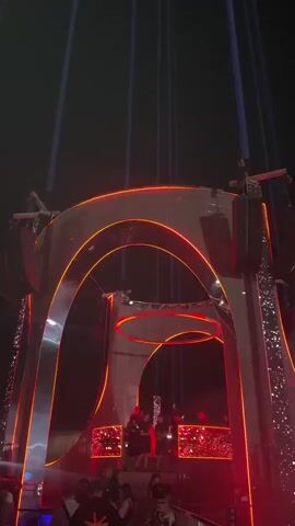 The video shows a dimly lit outdoor scene at night with a large, elaborate stage structure. The stage is illuminated with vibrant red and orange lights, and features a prominent arched entryway with mirrored accents. Several large speakers are mounted on the structure. In the background, thin beams of light are projected upwards into the dark sky. On the stage, several figures, likely performers or DJs, are visible behind what appears to be a DJ booth or console. The crowd is not clearly visible, but the presence of people suggests an event or concert. The overall atmosphere is energetic and festive, typical of a nighttime music performance.