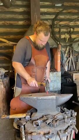 A man with a beard and hair pulled back in a bun, wearing a leather apron over a t-shirt, is in a workshop. He is standing over an anvil, holding a hammer in his right hand and a piece of glowing red metal with tongs in his left hand. He is actively striking the metal with the hammer. The workshop has wooden walls, a brick fireplace in the background, and various tools, including hammers and tongs, are visible on and around the anvil. The lighting suggests it is daytime.