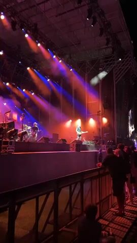 The video shows a live music performance on an outdoor stage at night. The stage is illuminated by purple and orange spotlights, creating a dramatic atmosphere. Several musicians are visible on stage, including a guitarist and a drummer. A crowd of people is gathered in front of the stage, watching the performance. The overall scene is one of a lively and engaging concert.