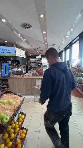 A man wearing a dark blue hoodie and dark pants is walking away from the camera inside a store. He is walking towards a counter where another person appears to be working. In the foreground, there are baskets filled with apples and lemons. Behind the counter, there are shelves stocked with various food and drink items. Large windows in the background reveal an outdoor area, possibly a gas station. The store appears to be well-lit with fluorescent lights on the ceiling and displays of products on shelves and in refrigerated units.