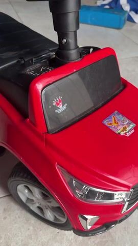 This media file shows a close-up, slightly shaky, indoor shot of a red toy car, likely for a child. The car is on a tiled floor, and the background is blurred, but appears to be a room with some household items, including a blue object and a bottle.

The toy car resembles a miniature SUV, with a black steering wheel and seat. There are some decals on the car: a red heart with a hand inside and the text "INDONESIA" is visible on the windshield. A colorful sticker, possibly featuring animals or a cartoon scene, is placed on the hood. The car's headlights and grill are detailed, mimicking a real vehicle. One of the wheels is clearly visible, showing a silver rim.

The lighting suggests it is daytime, but the specific time of day and weather conditions are not discernible. There are no people visible in the media file, and no particular activity or event is taking place other than the static presentation of the toy car. The overall impression is one of a child's toy, likely indoors in a residential setting in Denasri Kulon, Indonesia, given the "INDONESIA" text.