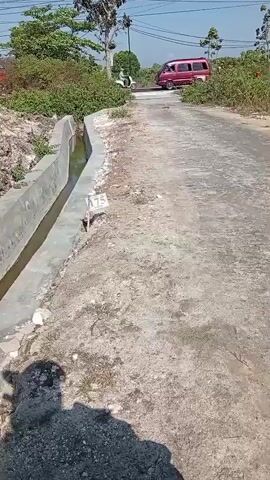 The video captures a slightly elevated view of a rural road on a sunny day. The road itself appears to be asphalt, with some rough patches and gravel on the sides. To the left of the road, there is a newly constructed concrete drainage ditch, its bottom filled with shallow, murky water. A small white stake with the number "75" is visible near the edge of the ditch.

The surroundings are characterized by sparse vegetation and some trees. On the far left, behind the ditch, a red car is partially visible. In the distance, at the top of a slight incline on the road, a person on a white scooter is moving away from the camera. Overhead, power lines crisscross the sky. The shadow of the person filming is cast prominently in the foreground on the right side of the frame. The overall impression is of a quiet, undeveloped area.