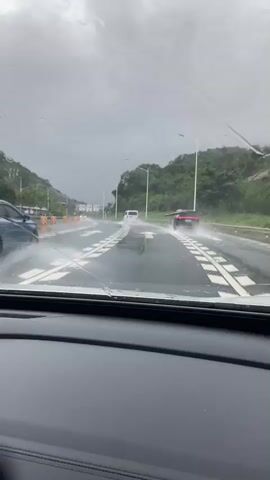 The video shows a view from inside a car on a rainy day. The windshield is covered in raindrops, obscuring the view of the road ahead. Several cars are driving on the wet road, with water splashing up around them. The road appears to be a multi-lane highway, with trees and hills visible on either side. The sky is overcast and grey. The windshield wipers are visible and appear to be in motion, attempting to clear the rain from the windshield.