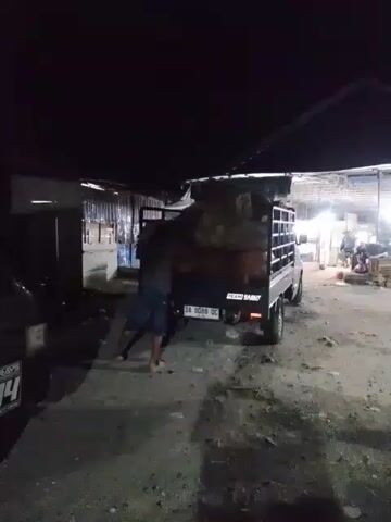 The video shows a nighttime scene outdoors with a truck. The truck bed is loaded with what appear to be large sacks or bundles. A person is seen pushing against the back of the truck, seemingly to move it. Another person, wearing a yellow vest, walks away from the truck toward the left side of the frame. The environment appears to be a poorly lit outdoor area, possibly a market or storage area, with some structures visible in the background. The ground is rough and uneven.