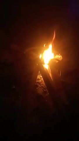The video shows a small campfire burning in the dark. Three pieces of wood are arranged to support the flames.  The fire is bright orange and yellow, illuminating the immediate area.  The surroundings are mostly black, indicating it is night.