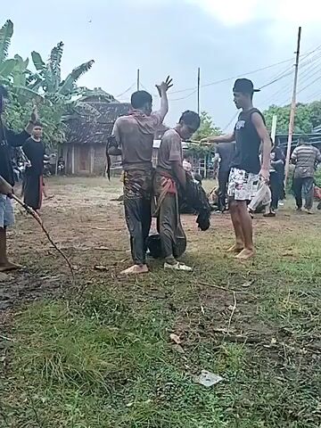 The video depicts a group of people gathered in a muddy field under an overcast sky.  Four individuals are in the foreground: one is wielding a long, thin whip; two others are standing close together, appearing to be participating in an activity of some kind; and the fourth person stands apart, watching.  Other people are visible in the background, some holding similar whips. A simple building is seen behind them, as well as trees and power lines.