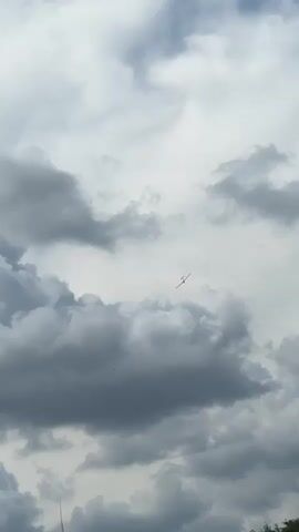 A distant, small white aircraft flies across a cloudy sky. The clouds are gray and white, with darker, heavier clouds below and lighter, wispy clouds above. The aircraft is a silhouette against the sky, its wings spread wide. The video captures the aircraft in flight for approximately five seconds.
