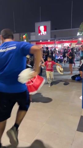 At night, a crowd of people is gathered on a brick-paved outdoor plaza. Some are participating in a dance, with a man in a blue t-shirt leading, twirling with a white cloth in his hand. To his right, a little girl in a red and white dress spins, and a small boy follows, wearing a sombrero. Nearby, a young woman in a black shirt and shorts dances. Many others can be seen dancing in the background. The area is decorated with flags and lights. In the backdrop are illuminated fast food restaurants, including KFC and Chili’s.