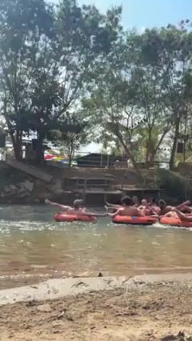 The video shows a group of people in inflatable tubes floating down a river. They are connected to each other, creating a line of tubes. The river flows through a natural environment with trees and vegetation along the banks. The sky is clear and sunny, suggesting a warm day. The camera is positioned on a sandy bank, providing a view of the river and the people enjoying the activity.