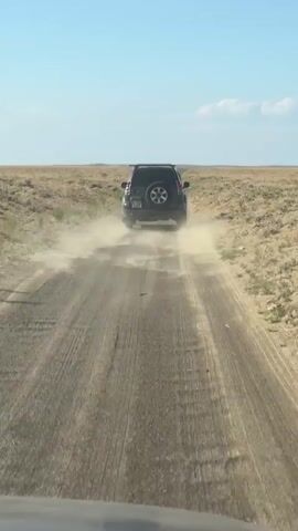 The video shows a black SUV driving away from the camera on a dusty, unpaved road in a dry, desolate landscape. The car is kicking up a large cloud of dust as it moves. The sky is clear and blue with a few wispy clouds. The ground on either side of the road is covered in dry, sparse vegetation. The camera appears to be mounted on another vehicle.