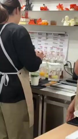 A woman is preparing a drink behind a bar, wearing a dark long-sleeve shirt and khaki pants, along with a beige apron with a white strap. She is adding an ingredient from a small bottle into a clear plastic cup filled with ice and a layer of white liquid at the bottom and a green layer on top. In the background, there are several bottles of clear liquids, a calendar, and Halloween decorations. Another person wearing an apron is visible to the right.