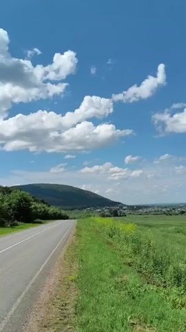 The video opens with a view of a paved road stretching into the distance, bordered by a vibrant green grassy field on the right. The road curves slightly to the left, leading towards a gentle, tree-covered hill. The sky is a brilliant blue, dotted with fluffy white cumulus clouds. As the video progresses, the camera pans to the right, revealing a vast expanse of green fields that extend to the horizon. In the distance, a small settlement with houses is visible. Further into the video, the landscape transitions, and a conical, rocky hill appears on the right side of the frame, silhouetted against the blue sky. The video captures the serene beauty of a rural landscape on a sunny day. There are no people, vehicles, or animals visible in the video.