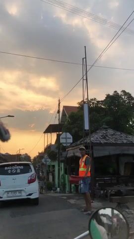 The media file depicts a street scene at dusk in Boro, Indonesia. The primary subjects are a man wearing a bright orange safety vest and a white car. The man is standing on the side of the road, holding a red flag or signaling device. He appears to be directing traffic or signaling to someone.

The scene is set on a paved road with buildings lining its sides. One building has a thatched roof and appears to be a small shop or dwelling. Another building, partially visible, has a red-tiled roof. The sky is filled with dramatic clouds, illuminated by the warm, golden hues of sunset, indicating the time of day. The weather appears to be clear with some cloud cover. Overhead, power lines are visible, a common sight in many urban and semi-urban areas.

A white car, possibly a hatchback, is parked on the left side of the road, facing away from the camera. On its rear window, there is visible text that reads "BAD QIENS" with a stylized symbol. The car's license plate is also visible, showing "H 1705 WD".

In the background, another person, a male, is walking away from the camera down the street. The overall mood suggested by the lighting and the presence of the traffic director is one of an ordinary evening in a bustling community, likely with ongoing daily activities. The focus on the safety vest and signaling device suggests a possible road maintenance or traffic management event. The presence of Indonesian writing on the car's license plate confirms the location context.