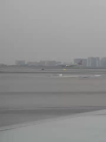 The video shows an airplane taking off on a runway. The airplane is white with a red tail and is speeding along the runway, which is gray. In the background, there are a number of tall buildings under a gray sky. A portion of the wing of the airplane in which the video is recorded is seen at the very bottom. The environment appears hazy.