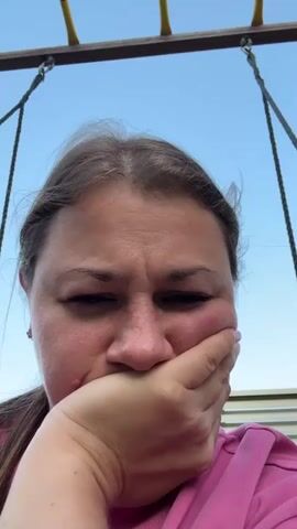 The video shows a close-up of a woman's face as she is on a swing set. She is holding her hand to her mouth, with her eyes wide open. The background is a blue sky.