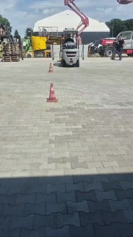 The video captures a forklift training session outdoors on a sunny day. The scene is an open paved area, likely a training ground or industrial yard, with a large white building in the background. A small, silver forklift with a driver is the central focus, navigating around two orange traffic cones placed on the grey interlocking brick pavement.

Other heavy machinery is visible in the background, including a yellow excavator and a red boom lift, suggesting an industrial or construction equipment context. A person is seen walking to the right of the frame near a large vehicle. The sky is blue with scattered clouds, indicating fair weather.

The forklift appears to be completing a maneuver, possibly part of a driving test or practice. The overall atmosphere seems to be one of focused activity and training. The time of day appears to be daytime, given the bright sunlight and shadows. There is no visible text in the frame.
