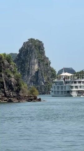 The video features a scenic daytime view of a large, white multi-decked cruise ship navigating through a bay. The ship, identified by "HERITAGE LINE" visible on its side, is moving between towering limestone karst islands covered in lush green vegetation. The water is calm and blue, reflecting the clear, bright sky. People can be seen on the upper decks of the ship as it moves past the striking rock formations. The scene depicts a peaceful journey through a picturesque landscape, likely in Ha Long Bay, Vietnam.